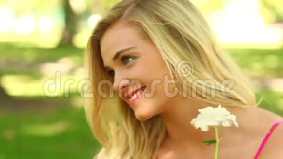 漂亮的金发女郎在公园里放松一边对着镜头微笑一边闻着一朵花视频的预览图