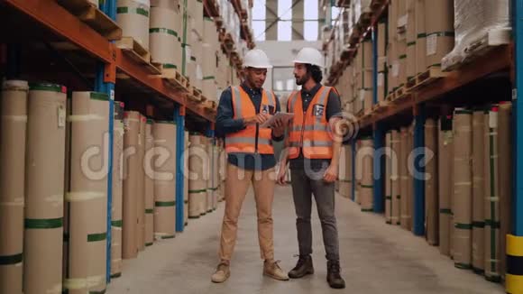 两名年轻男工人站在配送仓库身穿白色硬帽和反光夹克检查库存视频的预览图