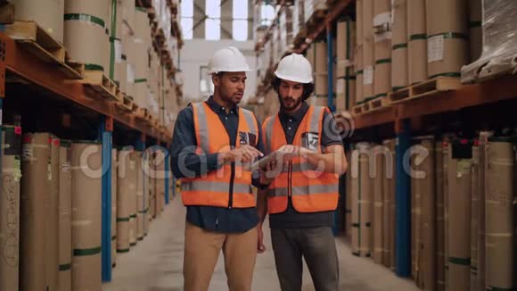 两名年轻的男性仓库工人穿着白色硬帽和反光背心使用数字平板电脑进行大规模分发视频的预览图