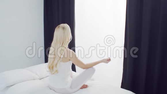 年轻女子在床上打坐漂亮的金发女孩做瑜伽运动早上在卧室里白天视频的预览图