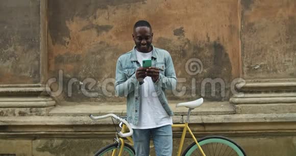 一位年轻的美国黑人男子穿着休闲服用智能手机站着俯身看着屏幕微笑着视频的预览图