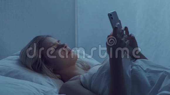 年轻的女人晚上躺在床上拿着智能手机美丽的金发女孩休息卧室里的暮色视频的预览图