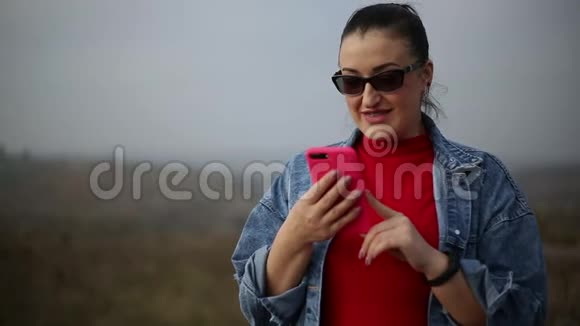 带有牙套的快乐女人的肖像使用粉红色智能手机戴太阳镜红色毛衣和蓝色牛仔裤夹克视频的预览图