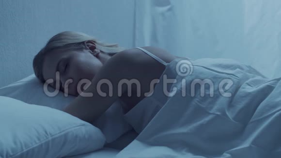 年轻的女人晚上躺在床上美丽的金发女郎卧室里的暮色窗外的月光视频的预览图