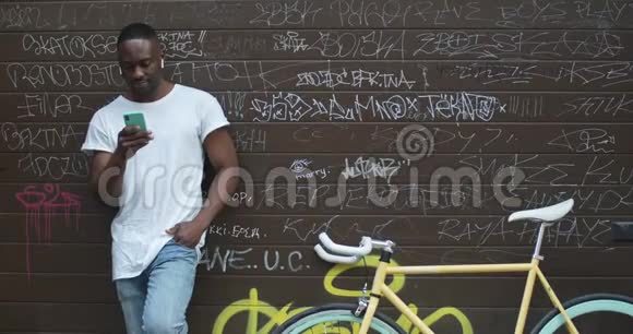 英俊的肌肉发达的非裔美国年轻人用他的智能手机站在时尚的现代自行车附近视频的预览图