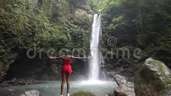 苗条的年轻女子穿着红色的浪漫在风景优美的瀑布前举手视频的预览图
