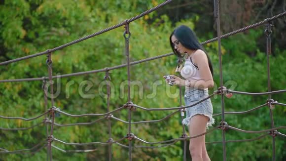 年轻的黑发女人戴着眼镜和短裤站在河上的吊桥上拍摄大自然视频的预览图
