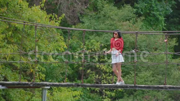 苗条的年轻黑发女人戴着粉红色的眼镜性感的红色衬衫带着电话站在河边的桥上看上去很远视频的预览图