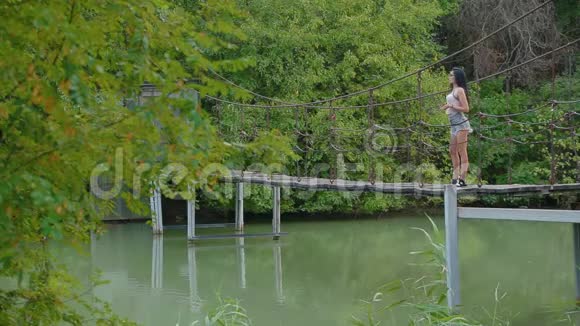 穿短裤的性感黑发女孩站在河边的吊桥上把石头扔进水里视频的预览图