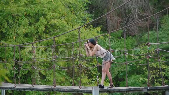 年轻的黑发女人戴着眼镜和短裤站在河上的吊桥上拍摄大自然视频的预览图