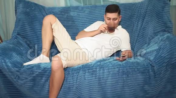 年轻人躺在沙发上看着电话喝着茶查看电子邮件互联网社交网络视频的预览图