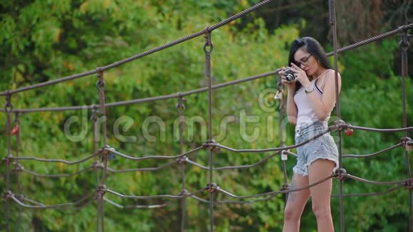 年轻的黑发女人戴着眼镜和短裤站在河上的吊桥上拍摄大自然视频的预览图