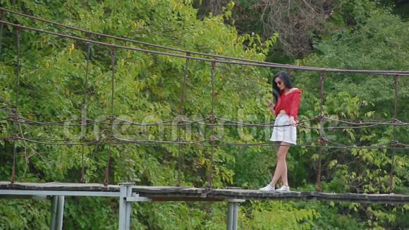 苗条的年轻黑发女人戴着粉红色的眼镜性感的红色衬衫带着电话站在河边的桥上看上去很远视频的预览图