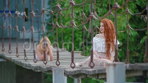 红色头发的女人坐在河上的吊桥上摆姿势模糊的背景摄影师为她拍照视频的预览图
