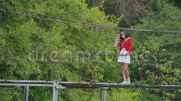 苗条的年轻黑发女人戴着粉红色的眼镜性感的红色衬衫带着电话站在河边的桥上看上去很远视频的预览图