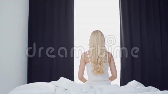 床上的年轻女子美丽的金发女孩醒来早上在卧室窗外的日光健康和休息视频的预览图