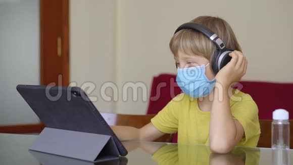 一个戴着防护面罩的小男孩在隔离期间坐在家里并在互联网学校使用平板电脑学习科罗纳视频的预览图