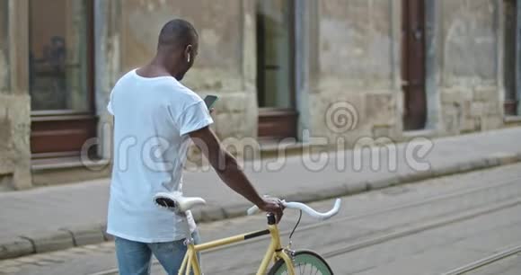 年轻的非裔美国人带着耳机用他的智能手机在老路上踩着时髦的现代自行车视频的预览图