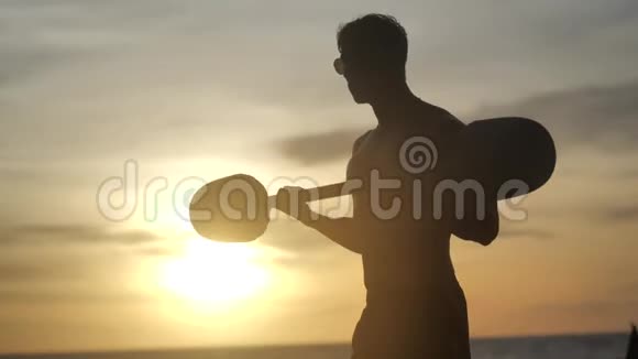 年轻的肌肉男在日落背景下在海滩上做杠铃举升运动员训练二头肌视频的预览图