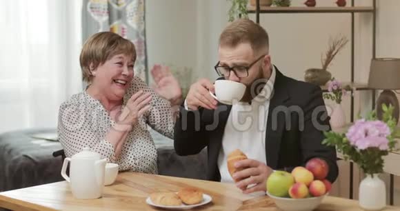 英俊的男人穿着闪闪发光的西装边吃边喝坐在母亲家的桌子上快乐的退休妇女视频的预览图