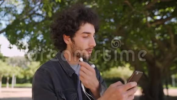 一位现代笑容可掬的年轻人在公园里用手机上网发短信把耳机插入耳朵的画像视频的预览图
