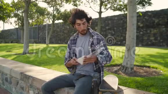 一个年轻人坐在滑板上吃三明治喝可重复使用的玻璃瓶里的水视频的预览图