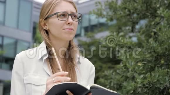 体贴的年轻女人在她的便笺上做笔记当她坐在户外的长凳上时看着远处视频的预览图