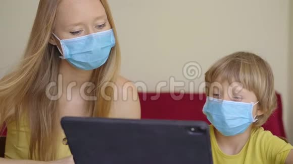 一位年轻的女士和她的小儿子在隔离期间坐在家里并在互联网学校使用平板电脑学习科罗纳视频的预览图
