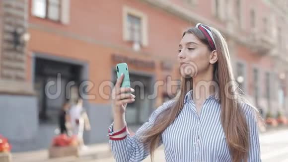 漂亮的年轻女孩有着棕色头发和时髦的头巾穿着条纹衬衫在她的智能手机上打视频电话视频的预览图