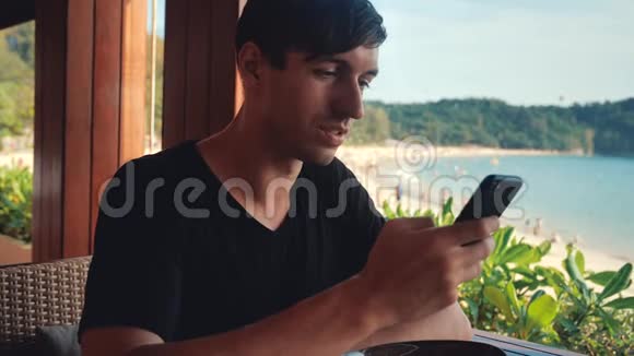 年轻的游客坐在海滩咖啡馆里在大海的背景上使用手机视频的预览图