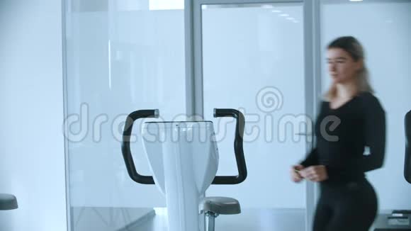 一位穿着黑色衣服的年轻女子坐在白色健身房的健身自行车上视频的预览图