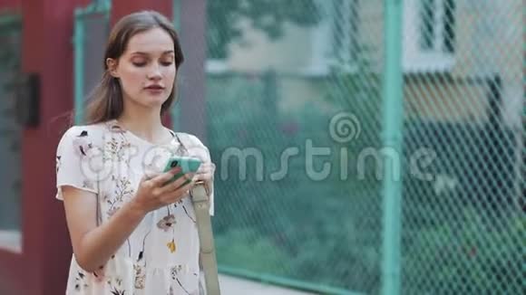 年轻漂亮的白种人棕色头发的女孩穿着白色的花裙用手握着她的智能手机把包放在肩上视频的预览图