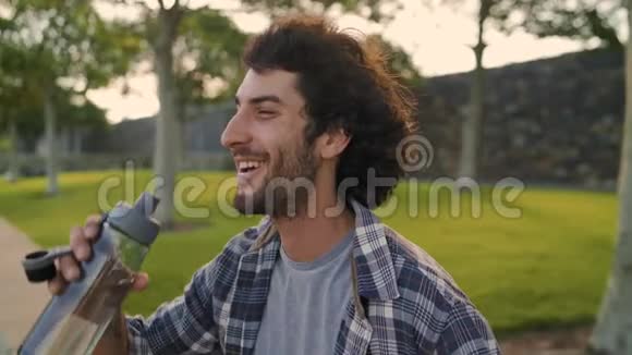 一位微笑的年轻人坐在公园里用可重复使用的水瓶喝水视频的预览图