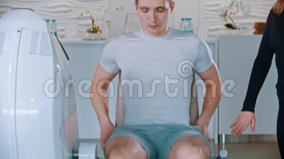 在健身房和健身教练一起训练一个男人坐在模拟器上做腿部训练他的女人视频的预览图