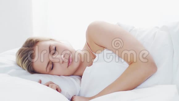 年轻的女人躺在床上拿着智能手机美丽的金发觉醒女孩早上在卧室里白天视频的预览图