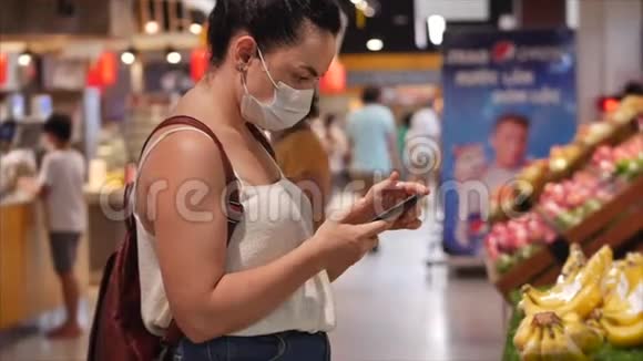 戴着冠状病毒流行面具的年轻女性在超市购物她的智能手机上的类型人们视频的预览图