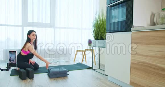 年轻女子正在做一个分裂伸展在瑜伽垫上在她的手机上录音视频的预览图
