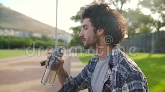 一个穿着休闲服的年轻人从公园里可重复使用的玻璃瓶里喝水的多利照片视频的预览图