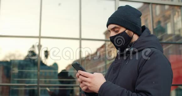 站在街上的人打字信息的侧面视图使用智能手机佩戴黑色棉防护面罩的男子视频的预览图