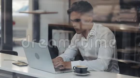 年轻的商人拿着笔记本电脑坐在桌子旁喝咖啡穿白衬衫漂亮的男经理视频的预览图