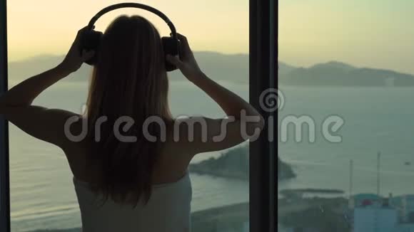 一位年轻女子在一间公寓里可以看到海岸和日出的全景她戴上无线耳机听着视频的预览图