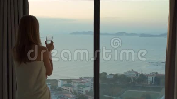 一位年轻女子在一间公寓里可以看到海岸和日出的全景她喝水视频的预览图