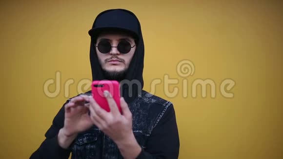 自信开朗的嬉皮士的肖像穿着全黑色使用带有粉红色外壳的智能手机背景是黄色的墙视频的预览图
