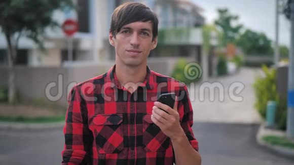 一张身着红色格子衬衫的英俊白人男子的肖像他站在城市郊区的户外用智能手机视频的预览图