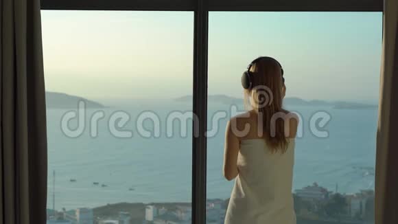 一位年轻女子在一间公寓里可以看到海岸和日出的全景她戴上无线耳机听着视频的预览图