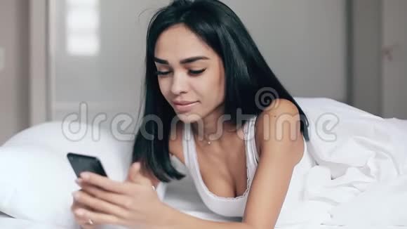 睡在床上的漂亮女孩看着智能手机上的信息微笑着视频的预览图