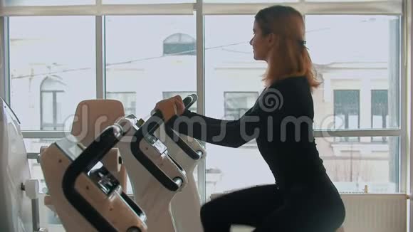 一位穿着黑色衣服的年轻女子在白色健身房的运动自行车上做腿上的运动视频的预览图