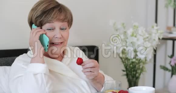 美丽的老妇人在床上吃早餐时说话和吃草莓使用智能手机的老太太视频的预览图