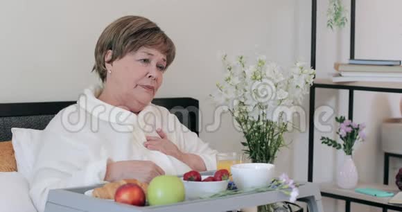 成熟漂亮的女士在床上吃早餐时心痛坐在家里的老年妇女感到不舒服视频的预览图