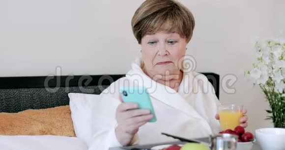 成熟的漂亮女人使用现代智能手机在床上吃早餐时微笑穿着家居的长者视频的预览图